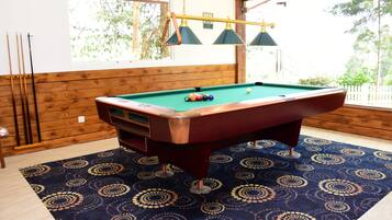 A pool table with balls, surrounded by a patterned carpet, wooden walls, and a window with a view of trees.
