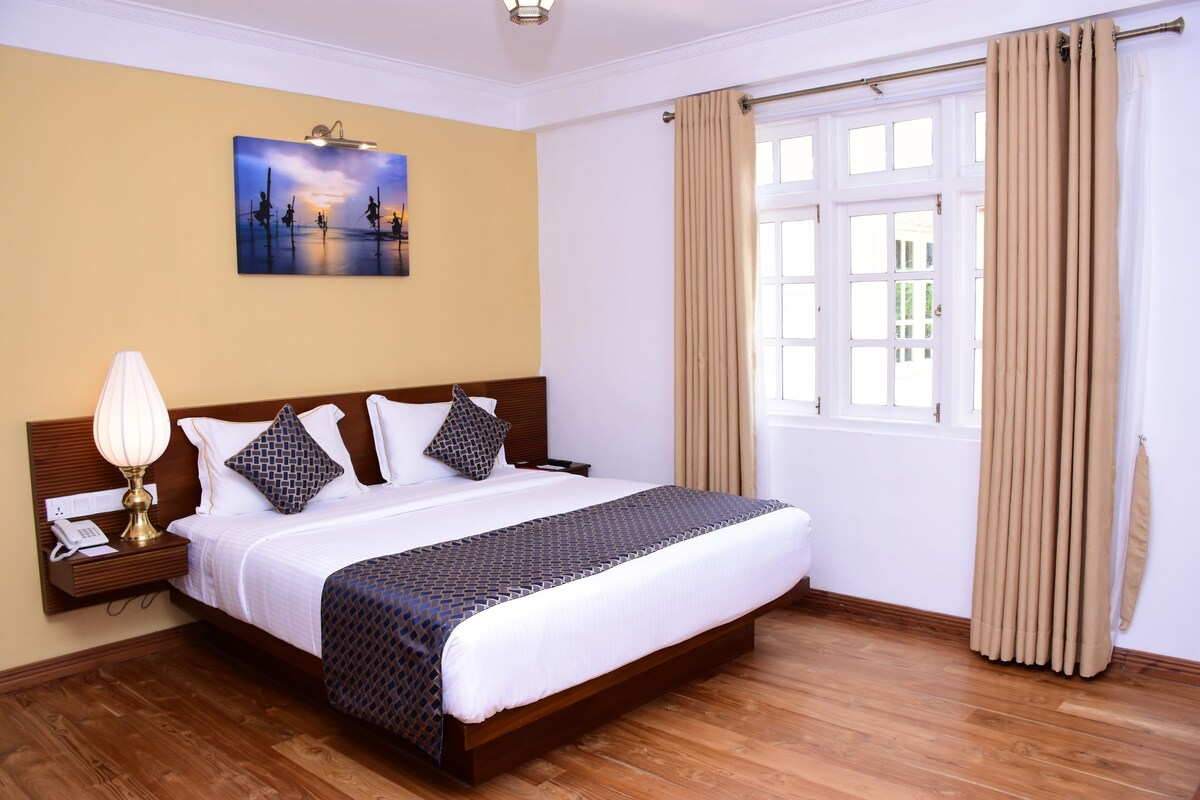 A hotel room with a large bed, a nightstand with a lamp, a painting on the wall, and a window with curtains.