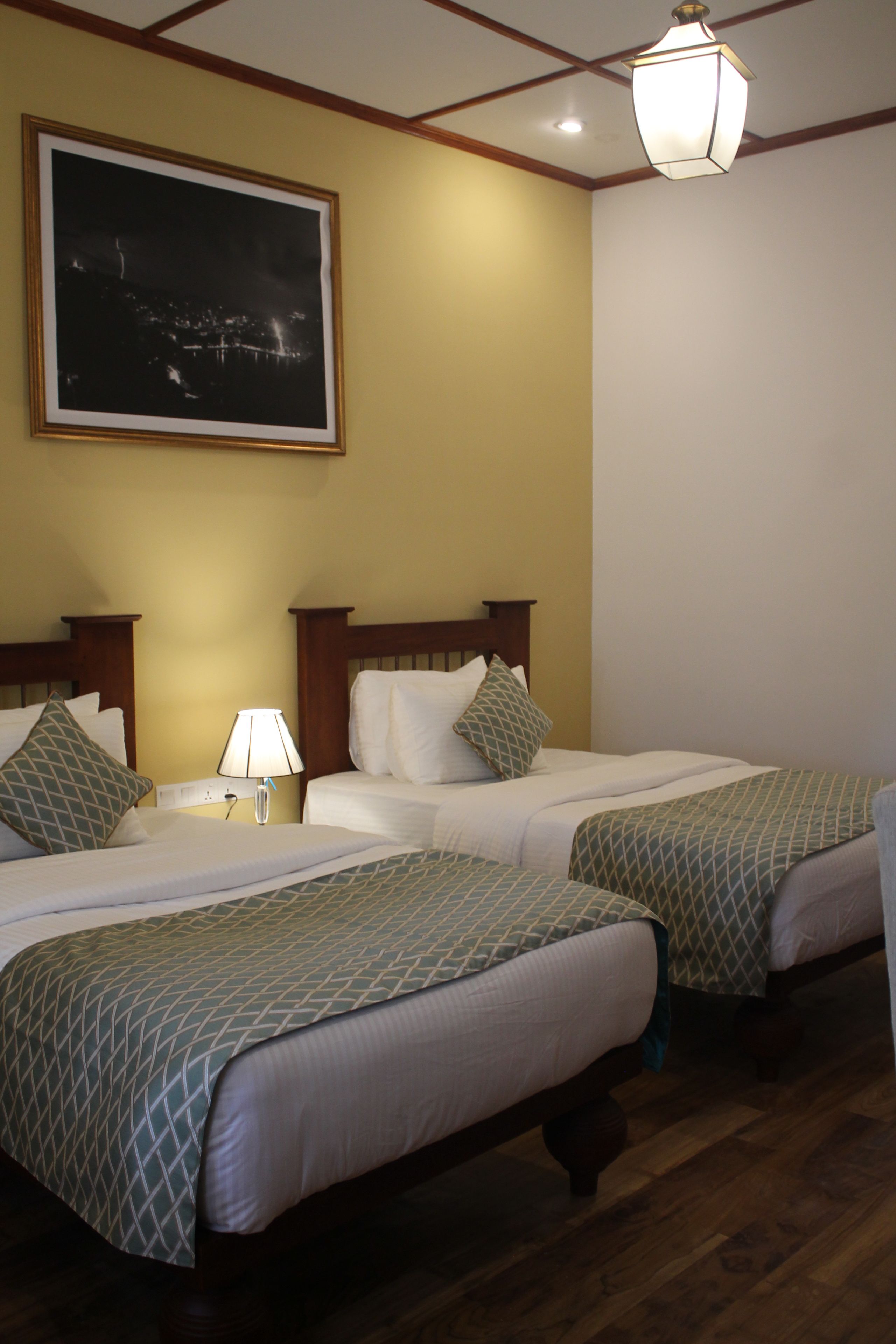 Habitación de hotel con dos camas, una mesita de noche con lámpara, un cuadro enmarcado en la pared y una lámpara colgante.