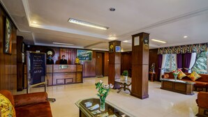 Un vestíbulo de hotel con mostrador de recepción, una zona de estar con sofá y sillones, un mostrador de recepción con un reloj y un jarrón con flores sobre una mesa.
