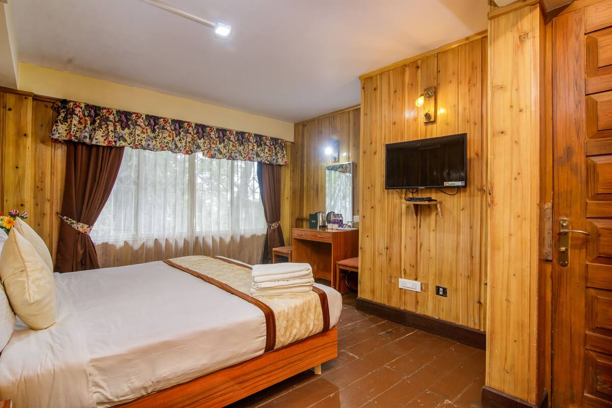Una habitación con paneles de madera, una cama, un escritorio, un televisor y un ventanal con cortinas.