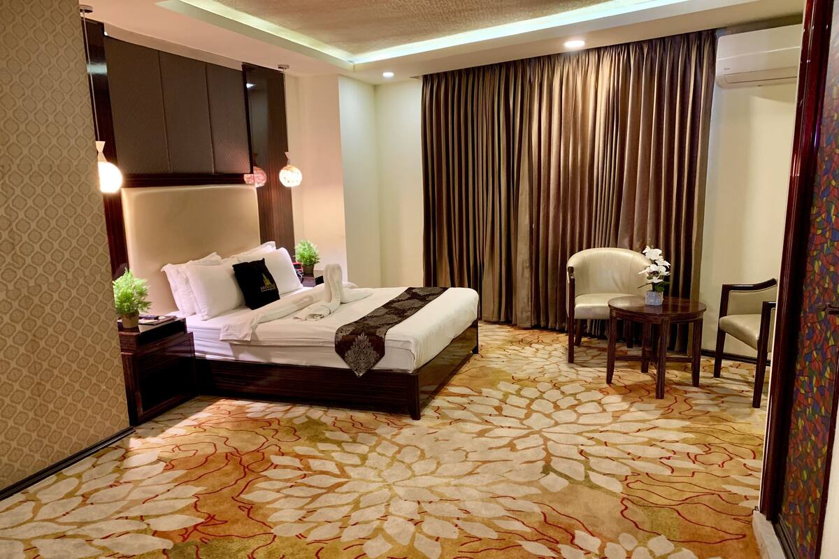 A hotel room with a large bed, a desk, a chair, and a large window with curtains.