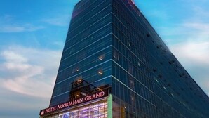 Un grand hôtel moderne, avec une façade en verre, sur lequel on peut lire en lettres bien visibles le nom « HOTEL NOORJAHAN GRAND ».