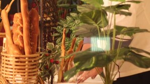 Un chef, vêtu d’un uniforme blanc et coiffé d’un chapeau, travaille derrière un comptoir où sont disposés des paniers de pain et des plantes en pot.