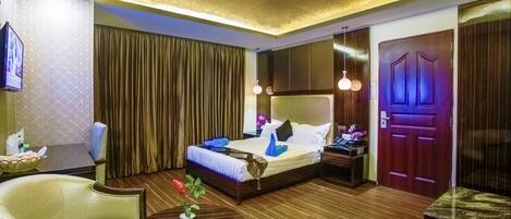 Une chambre d’hôtel avec un lit, une chaise, une petite table avec une plante, un plancher en bois et une porte avec une lumière violette.