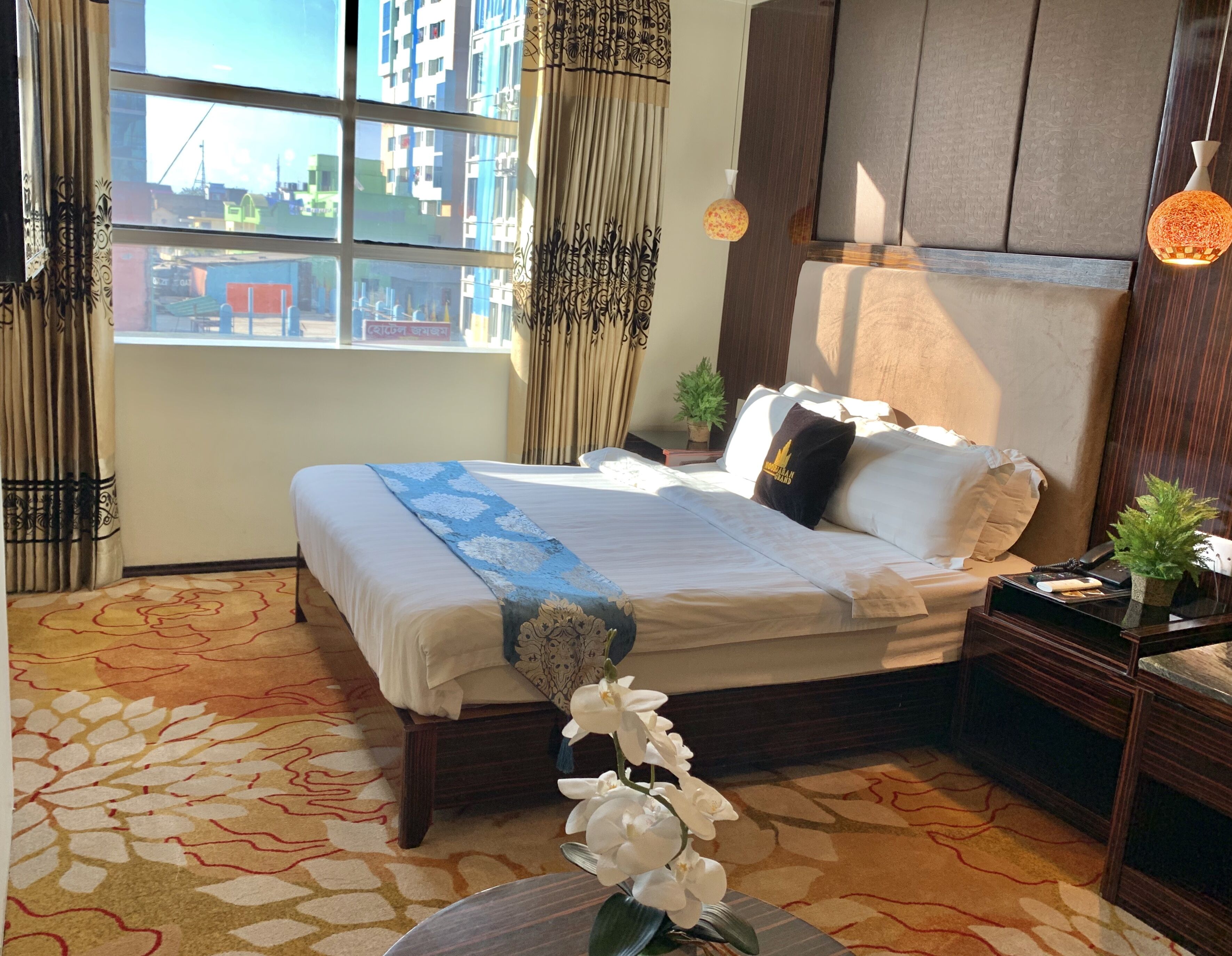 A hotel room with a large bed, a small table with a plant, a nightstand with a phone, and a view of buildings through the window.