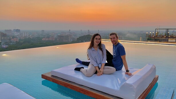 Un couple est assis sur un matelas, près d’une piscine à débordement, et profite de la vue sur la ville au coucher du soleil.