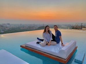 Una pareja sentada sobre un colchón junto a una piscina infinita con vista a la ciudad al atardecer.