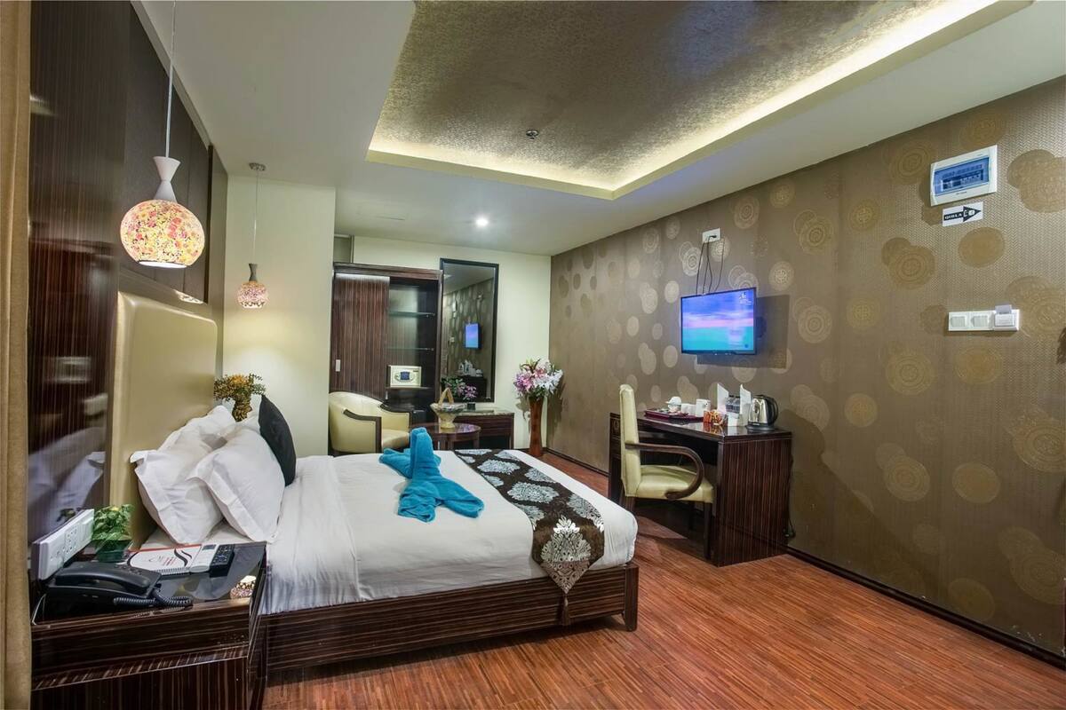 A hotel room with a bed, bedside table, TV, desk, chair, and a wall-mounted TV.