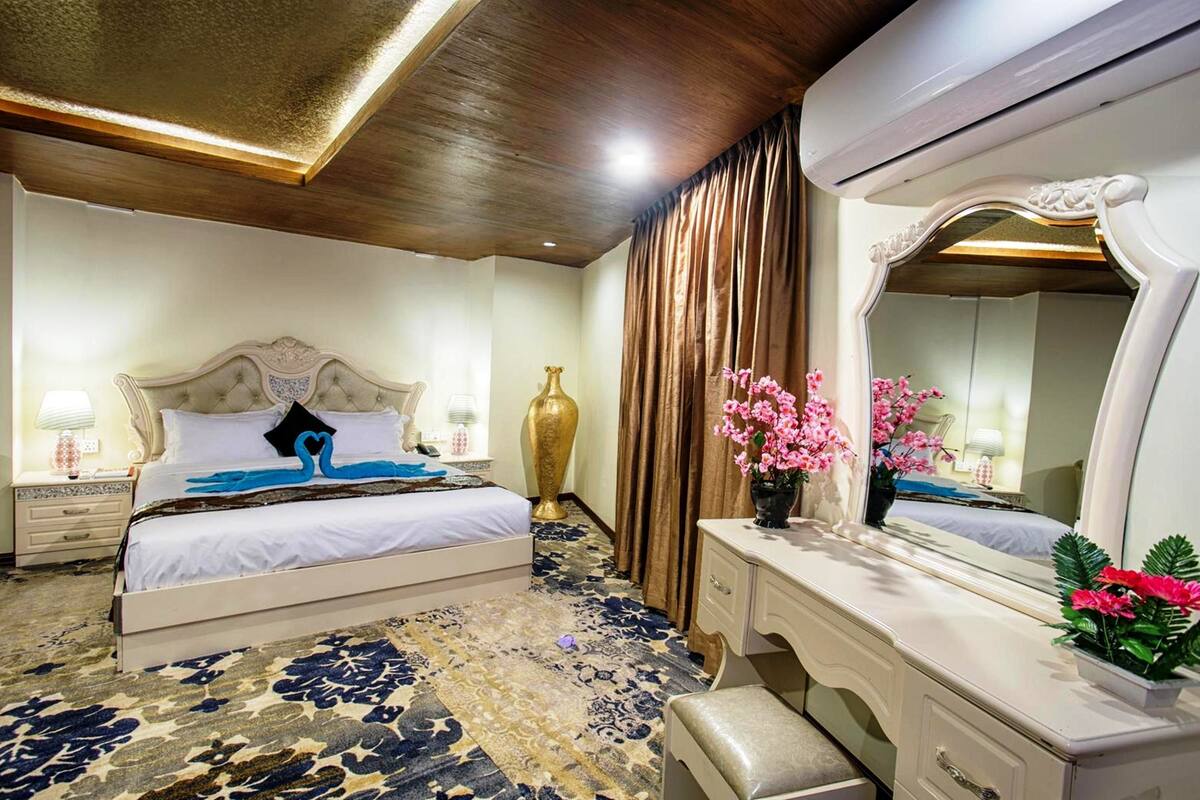 A hotel room with a bed, a vanity with a mirror, a nightstand, and a decorative vase.