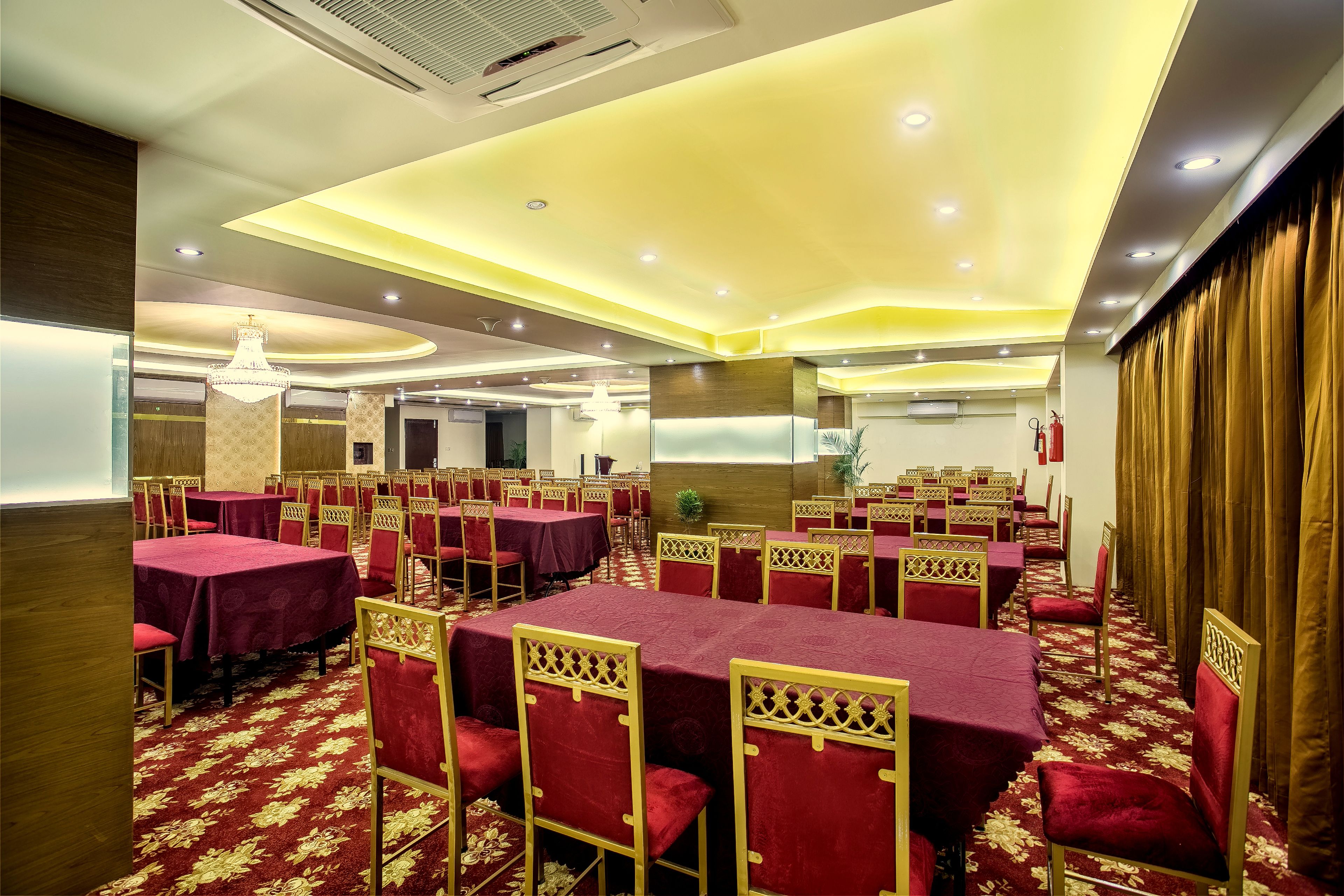 A banquet hall with tables covered in red tablecloths and gold chairs, arranged in rows.