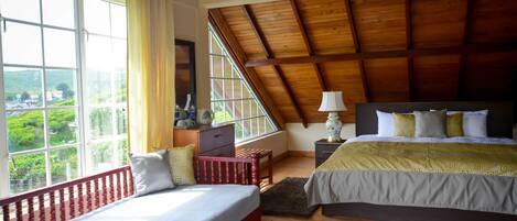 A bedroom with a large window, wooden ceiling beams, a bed with a patterned bedspread, and a nightstand with a lamp.