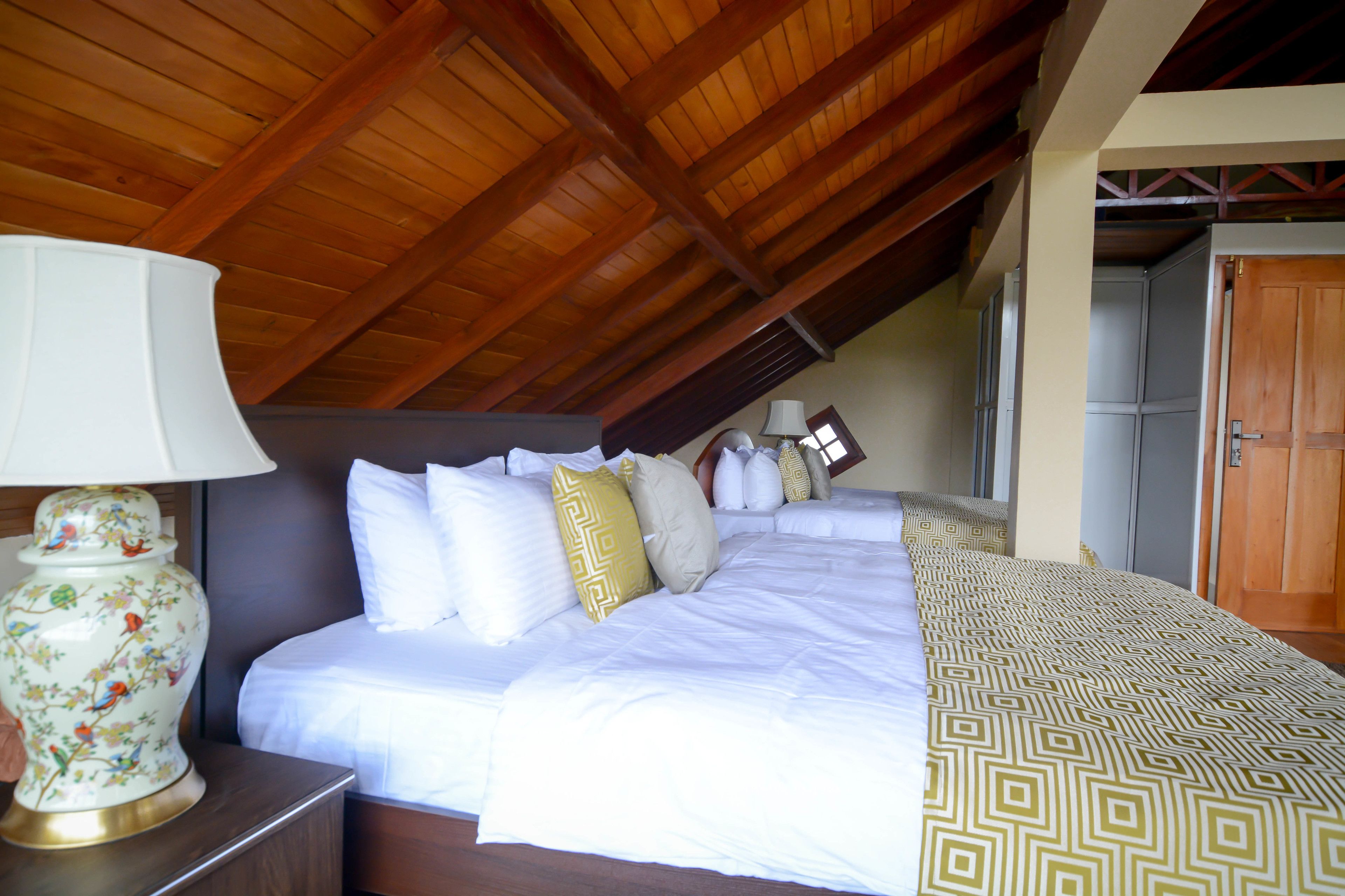 A neatly made bed with white bedding and decorative pillows, a wooden nightstand with a lamp, and a wooden ceiling.