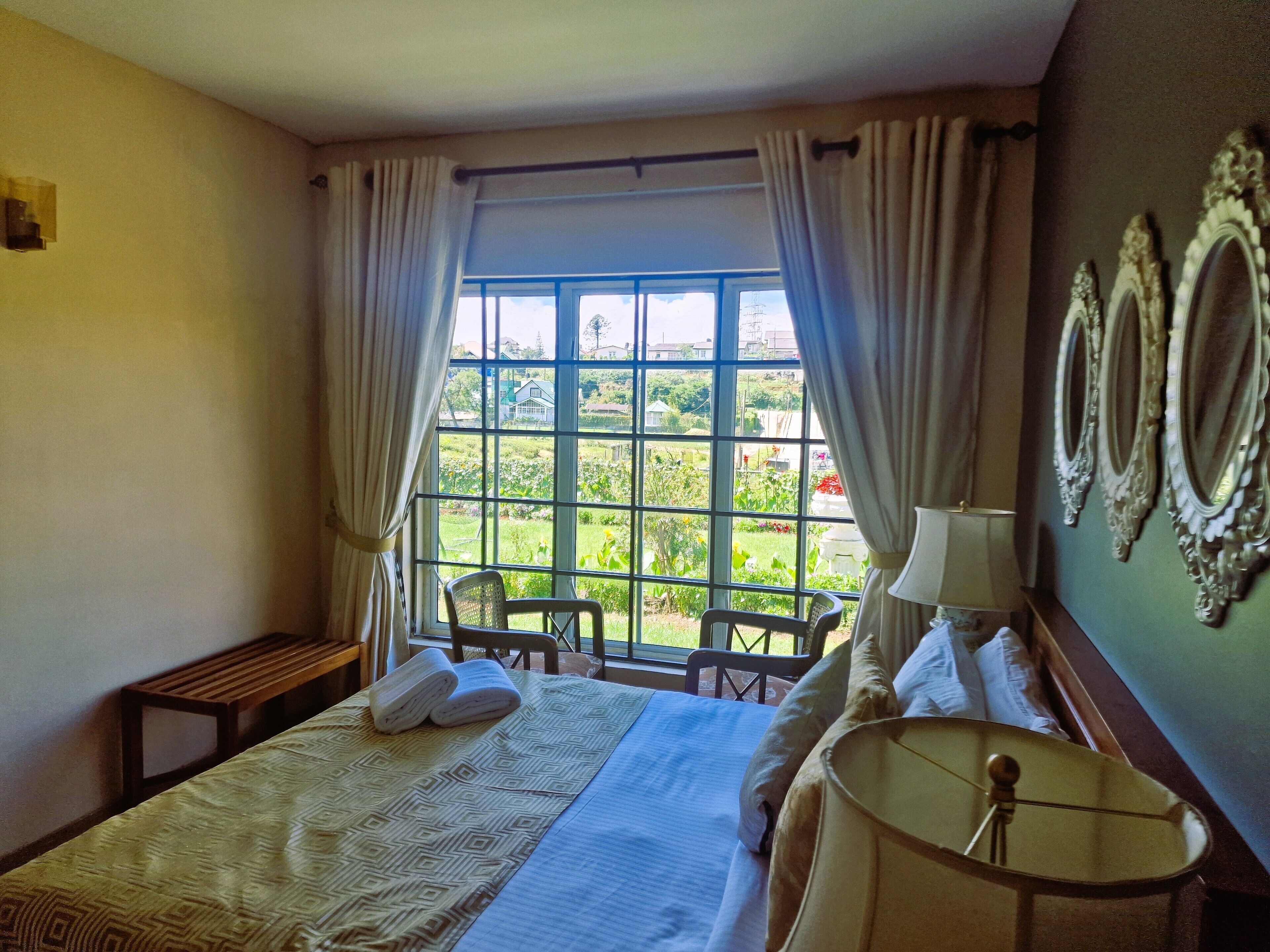 Une chambre d’hôtel avec une grande fenêtre, un lit recouvert d’une couvre-lit à motifs, une lampe de chevet et une chaise près de la fenêtre.