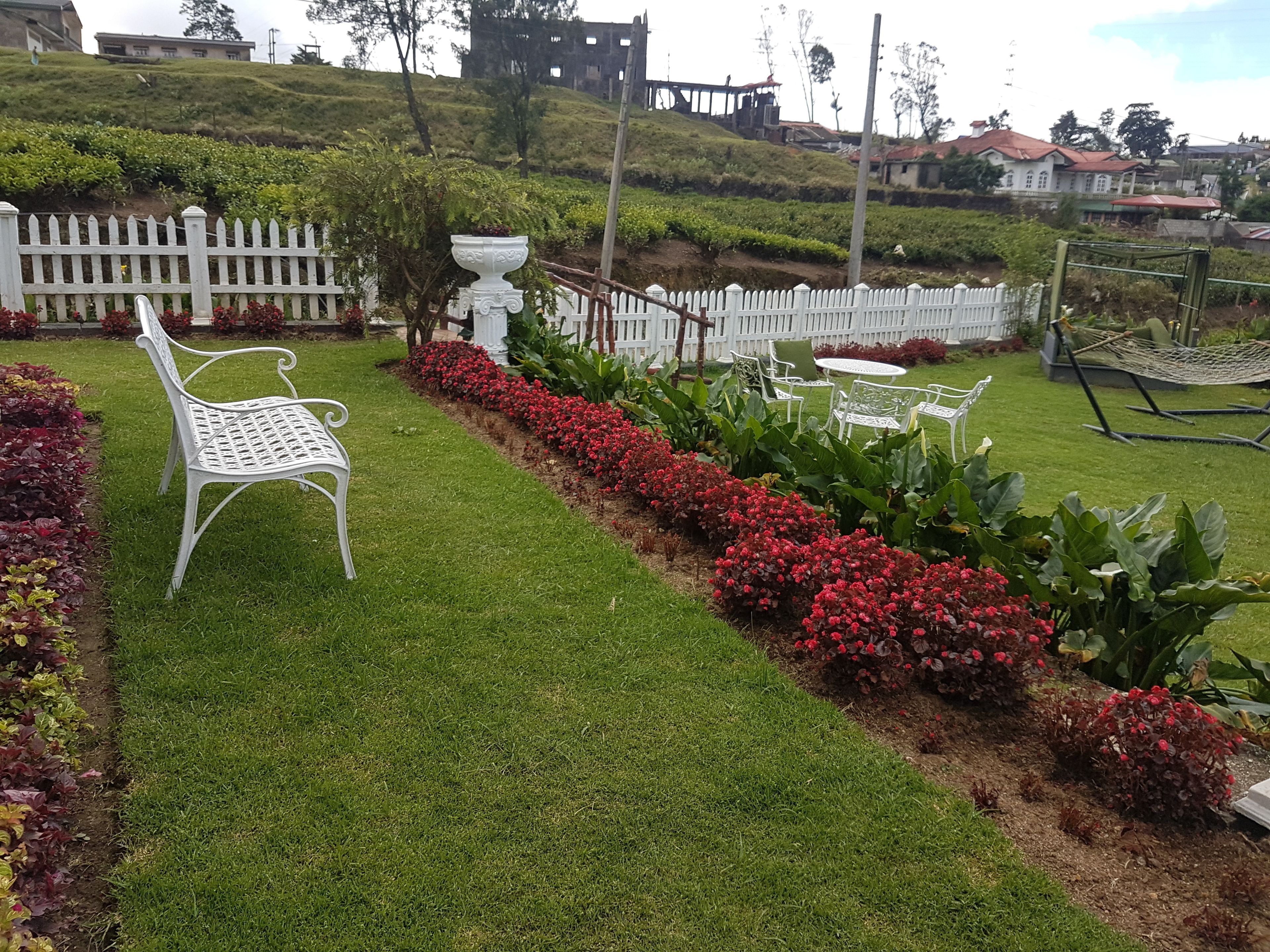 A well-maintained garden with a white metal bench, a white fence, and red flowering plants.