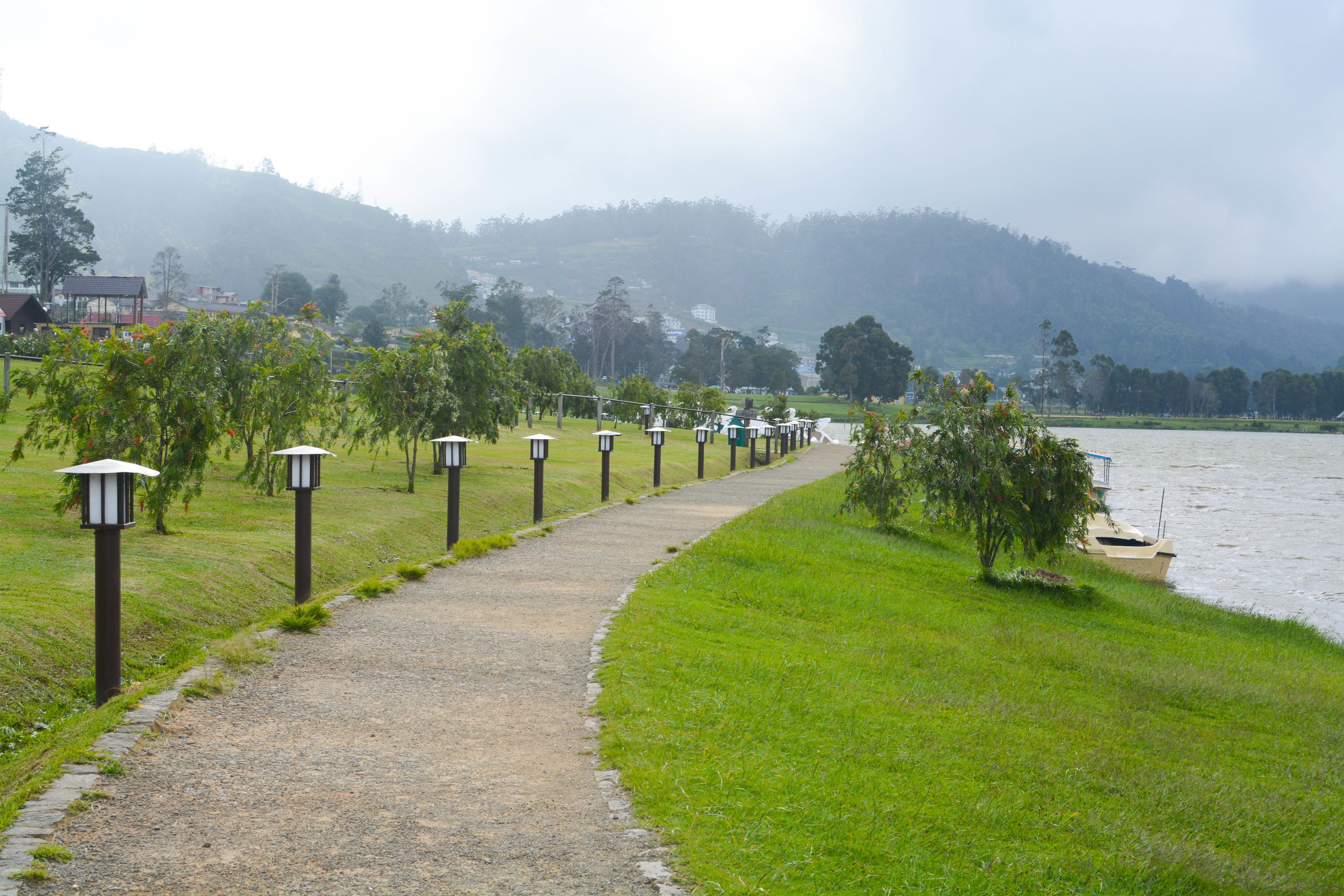 A gravel path alongside a body of water, flanked by lamp posts and greenery.