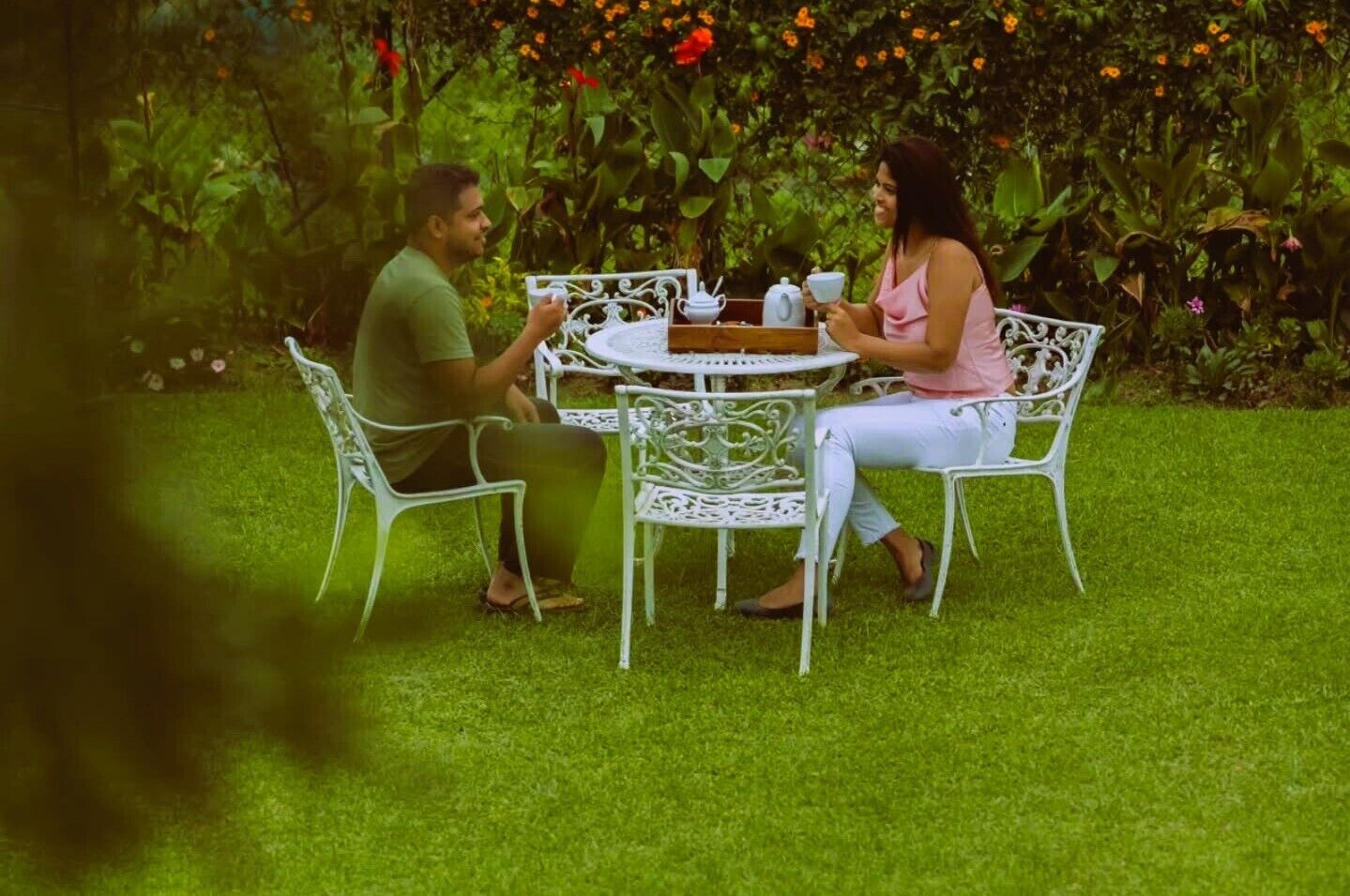 Two people sitting at a white metal table with chairs in a garden with green grass and flowering plants.