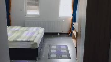 Quarto com uma cama, radiador, janela com persianas, porta e guarda-roupa.