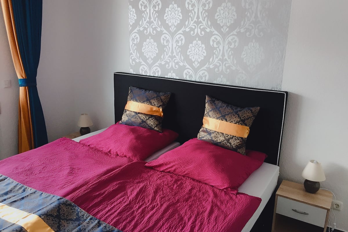 Een tweepersoonsbed met een bedsprei met patroon en twee decoratieve kussens, een nachtkastje met een lamp, en een muur met een decoratief motief.