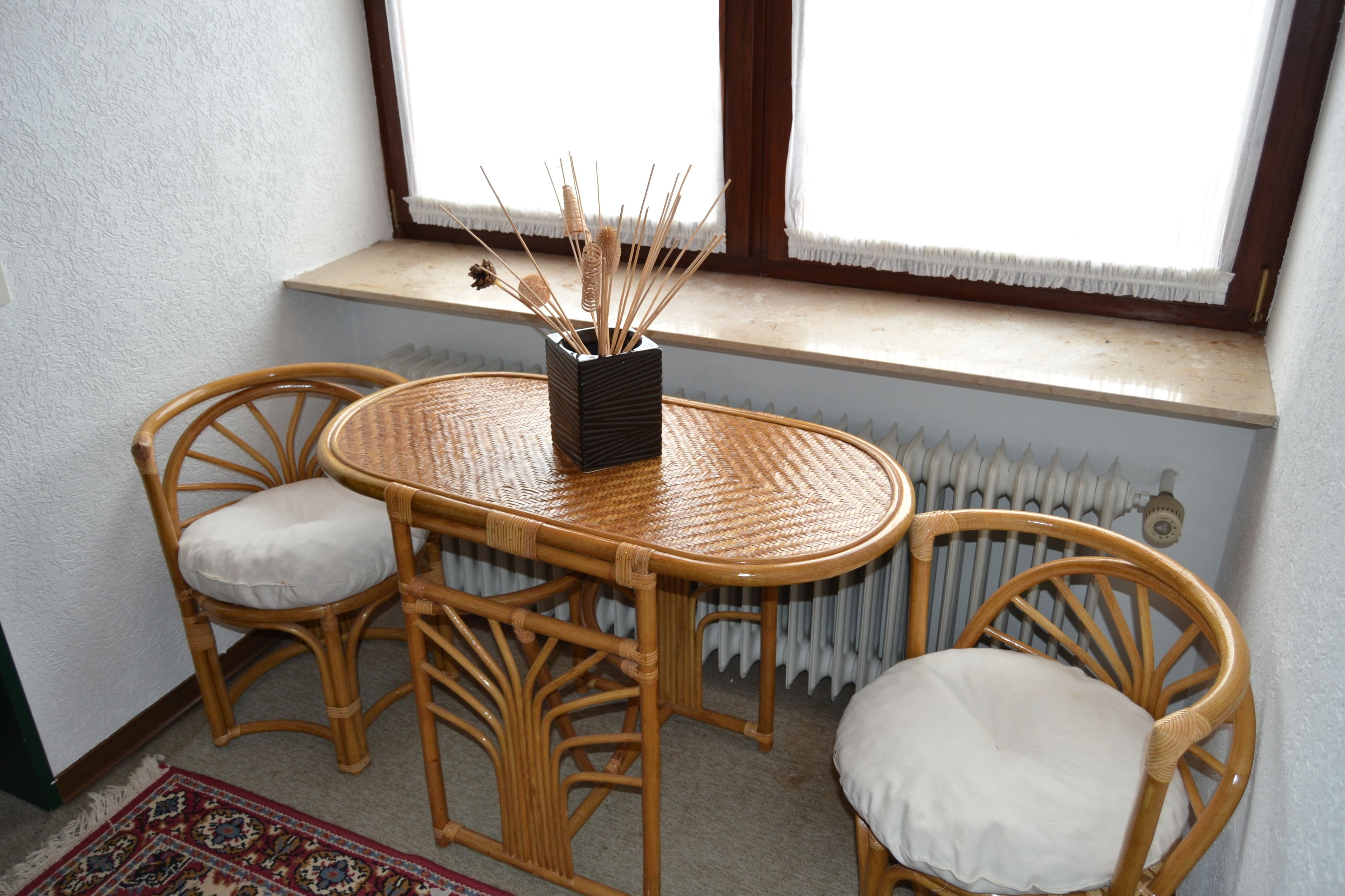 A corner room with a wicker table and two chairs, a small window with a view, and a radiator below.
