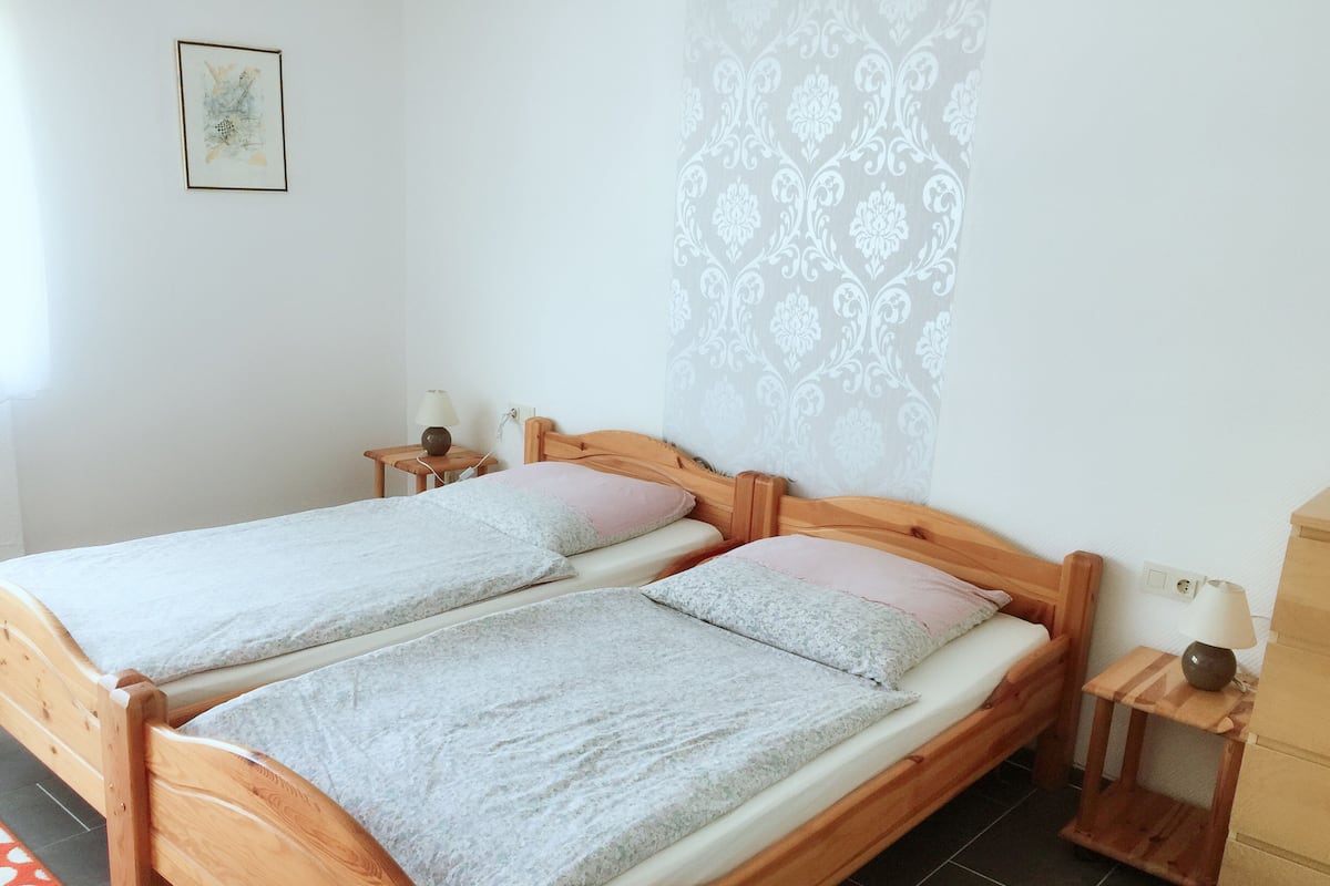 Een tweepersoonsbed met houten frame en twee kussens, een nachtkastje met lamp, en een muur met decoratief patroon.