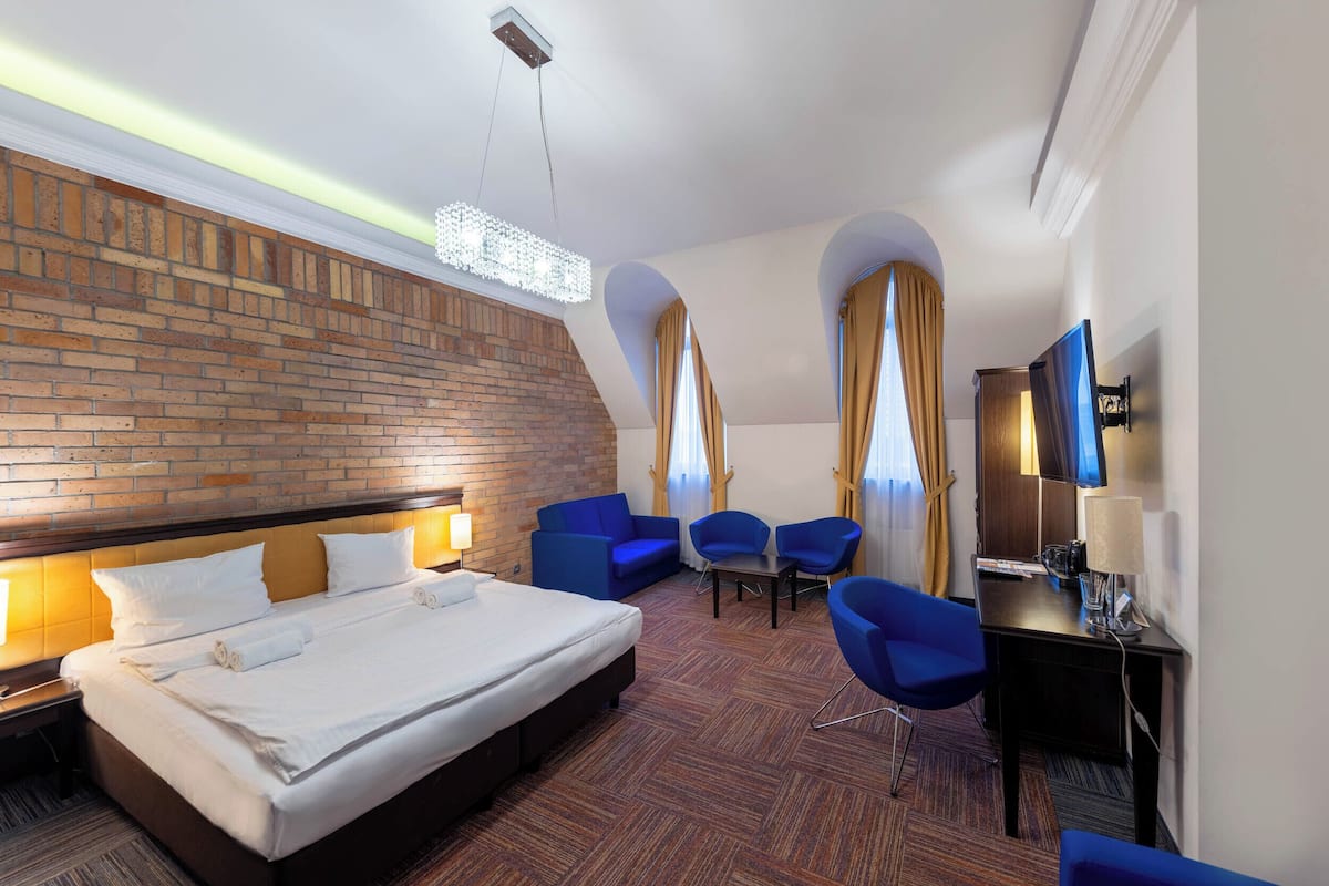 A hotel room with a large bed, two blue armchairs, a desk with a lamp, and a TV.