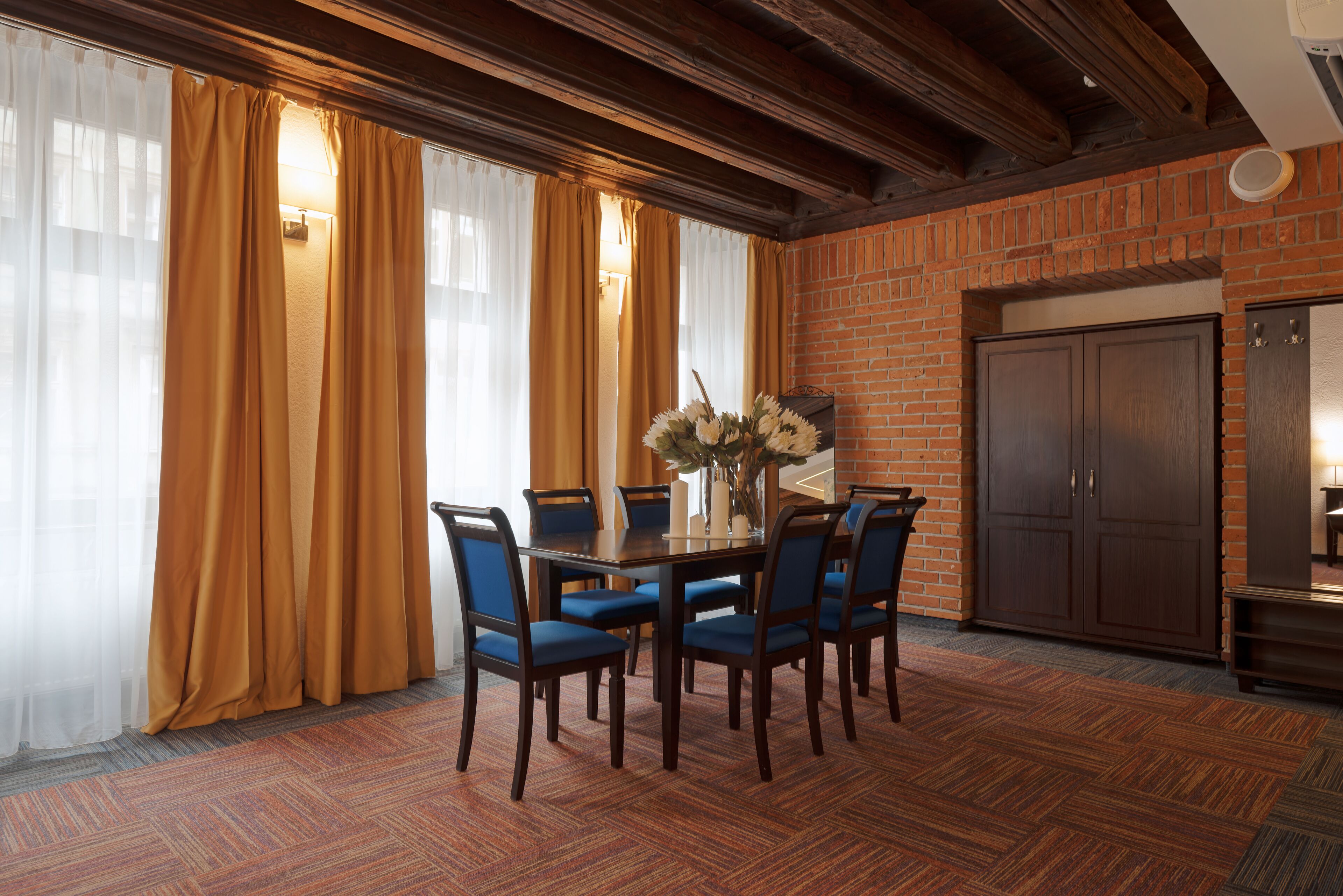 A dining area with a brick wall, wooden ceiling beams, and a table set for six with blue chairs.