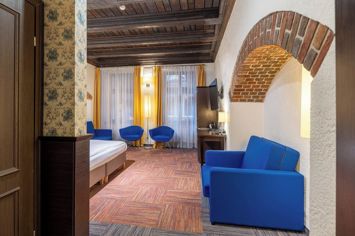 A hotel room with a bed, blue armchairs, a desk, a television, and a brick archway.