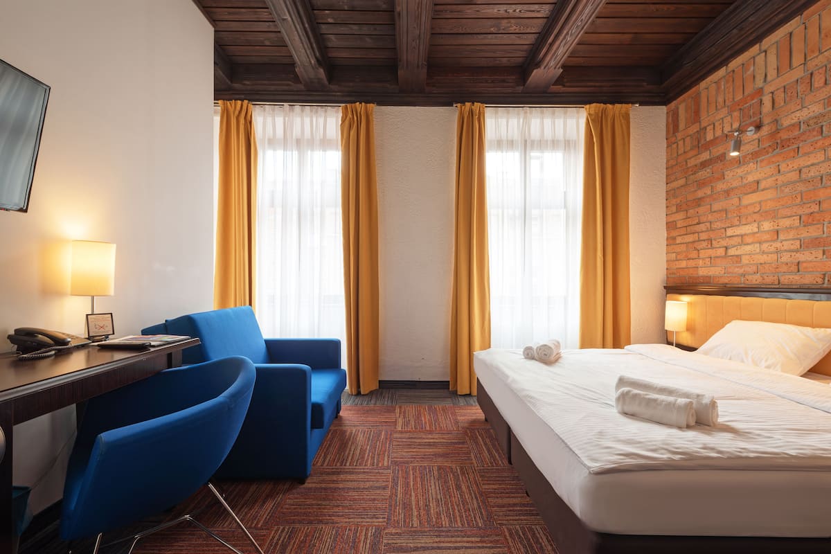 A hotel room with a bed, a desk, two blue chairs, and a brick wall.