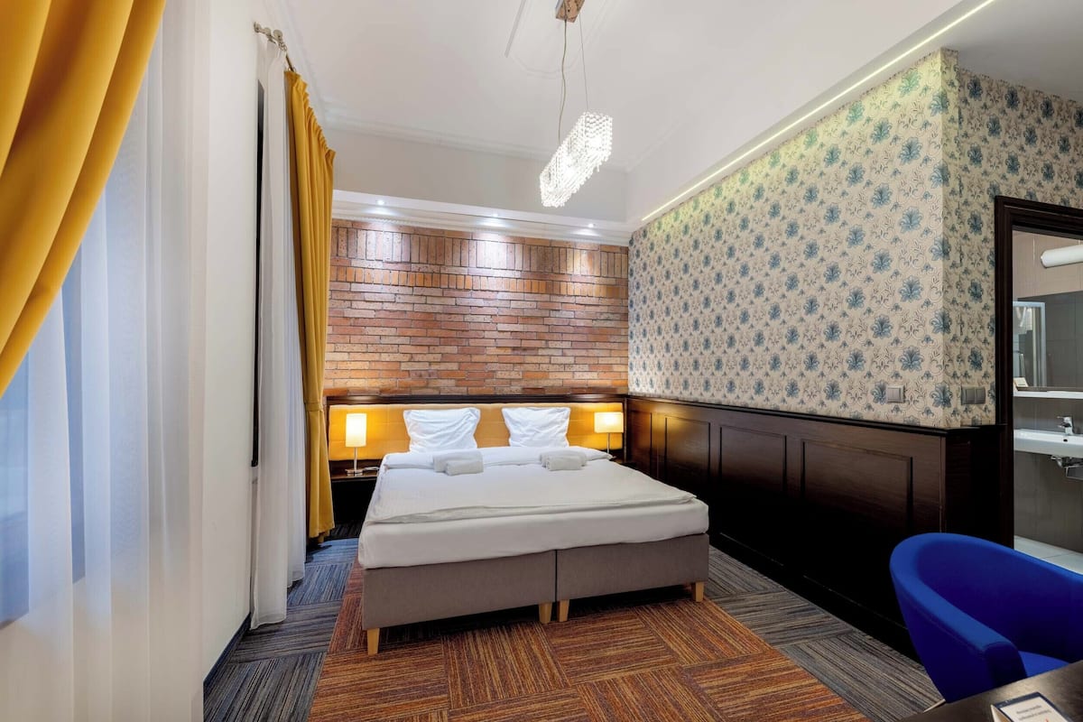 A hotel room with a bed, wooden headboard, patterned wallpaper, and a blue chair.