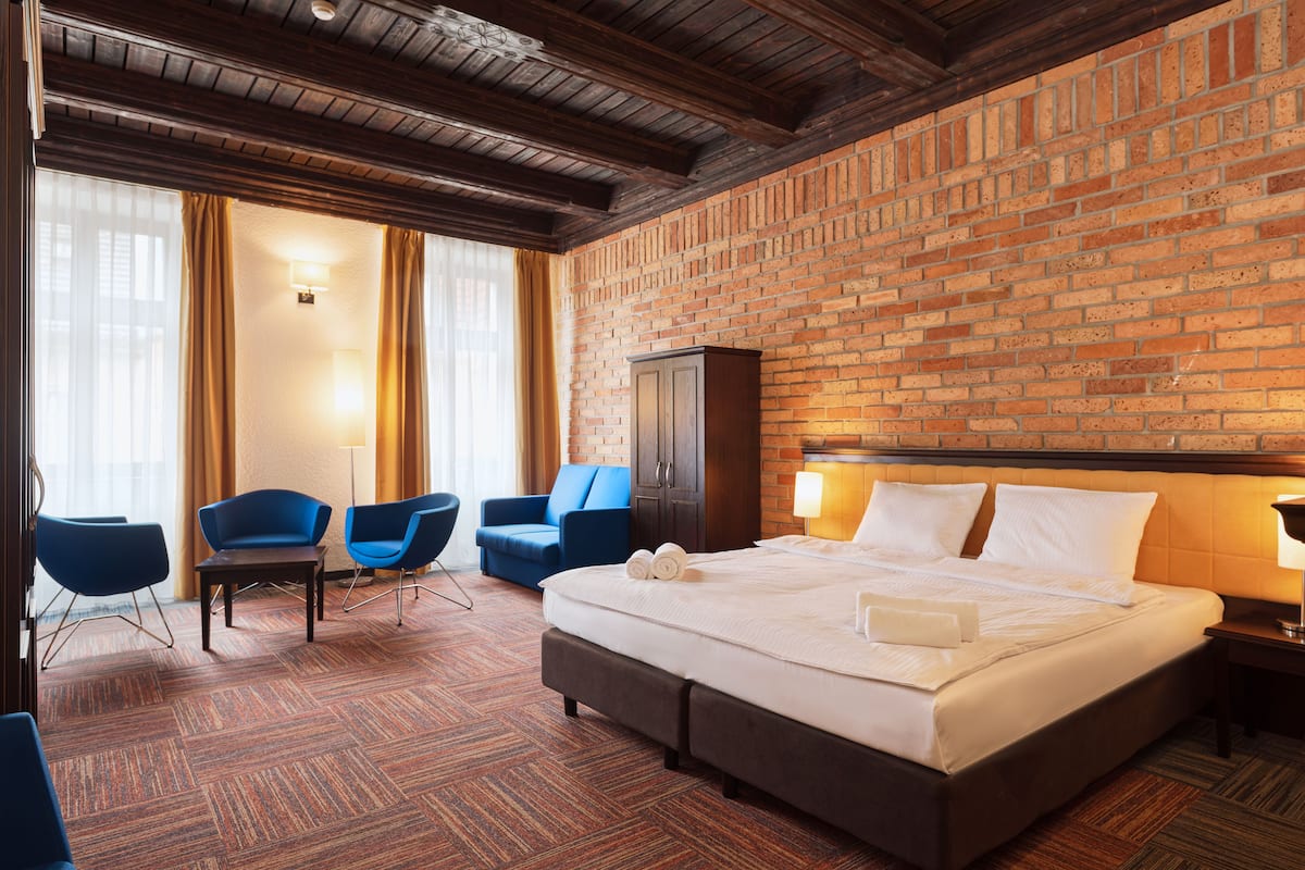 A hotel room with a brick wall, a large bed, two blue chairs, a wooden cabinet, and a carpeted floor.
