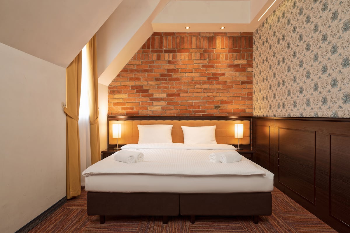 A hotel room with a brick-patterned headboard, a large bed with white linens, two bedside lamps, and a patterned wallpaper on the adjacent wall.