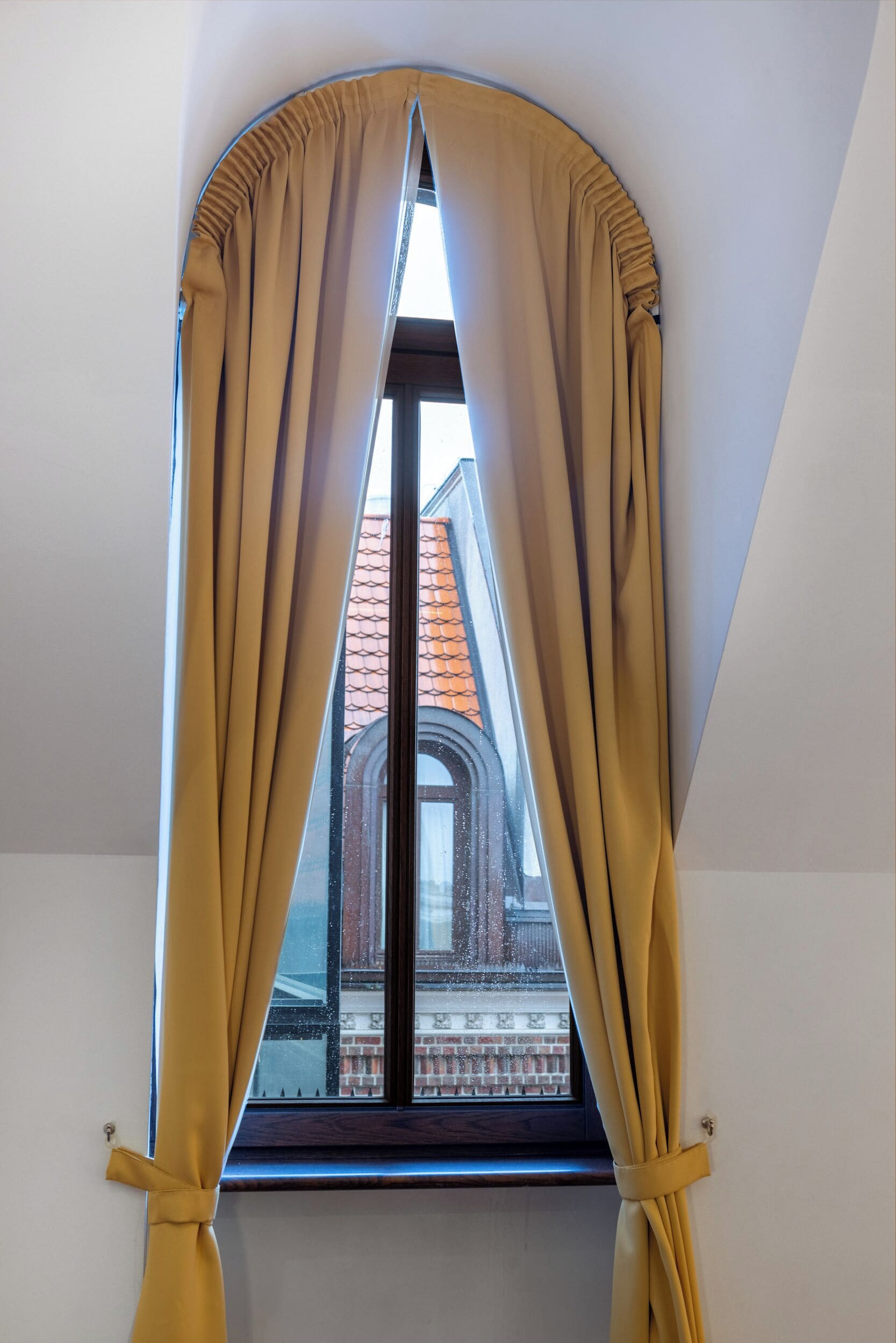 A window with yellow curtains tied at the bottom, revealing a view of a building with a red-tiled roof.