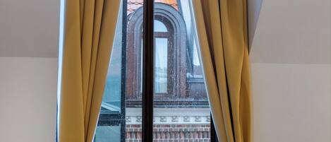 A window with yellow curtains tied at the bottom, revealing a view of a building with a red-tiled roof.