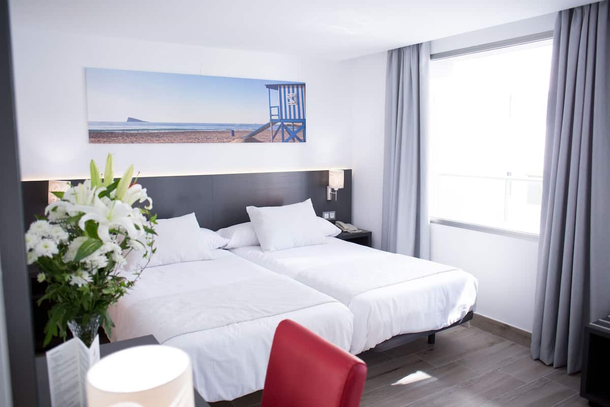 Habitación de hotel con dos camas, una mesita de noche, una lámpara, un florero con flores y un cuadro de una playa.