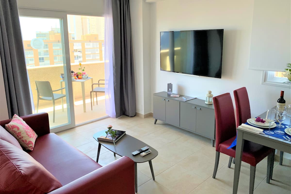 Una sala moderna con sofá, una mesa de centro, una mesa de comedor con sillas, un televisor montado en la pared y un balcón con mesa y sillas.
