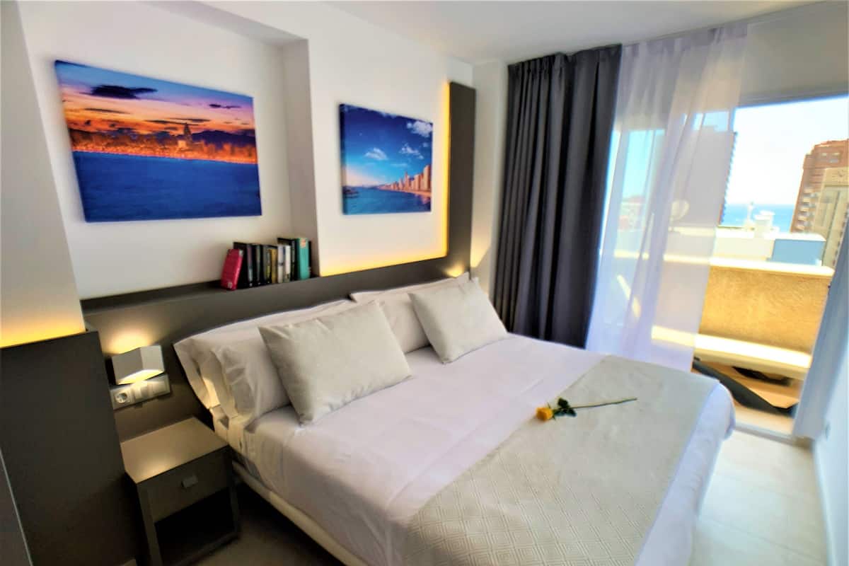 Habitación de hotel con una cama grande, dos cuadros en la pared, una mesita de noche y un ventanal con cortinas.