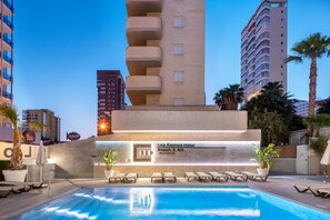 A hotel pool area with lounge chairs, a sign for Los Alamos Hotel Beach & Art, and a tall building in the background. - Hotel Los Alamos (Benidorm)