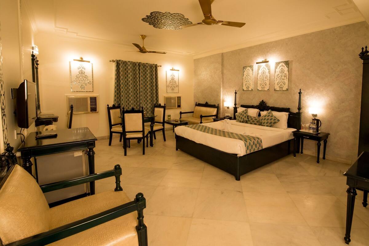 A spacious bedroom with a large bed, a desk, and a chair. There is a ceiling fan, a TV, and wall-mounted lights.