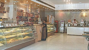 Una panadería con varios mostradores repletos de productos horneados, un mostrador con caja registradora y una zona de estar con mesa.