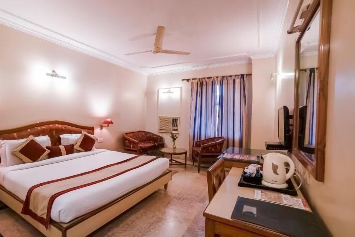 Quarto de hotel com cama, escrivaninha, cadeira, televisão, ventoinha no teto e janela com cortinas.