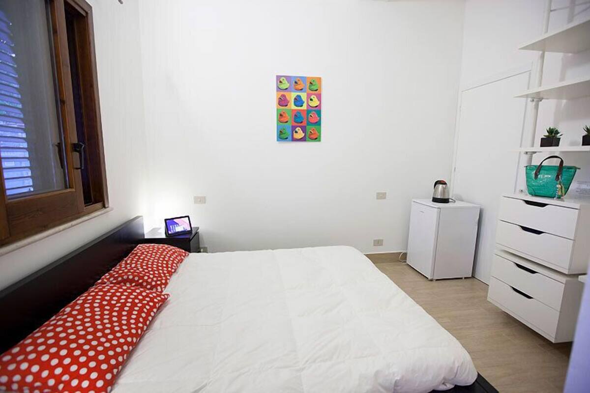 A bedroom with a bed, a small bedside table, a nightstand, a dresser, a window, and a wall-mounted artwork.
