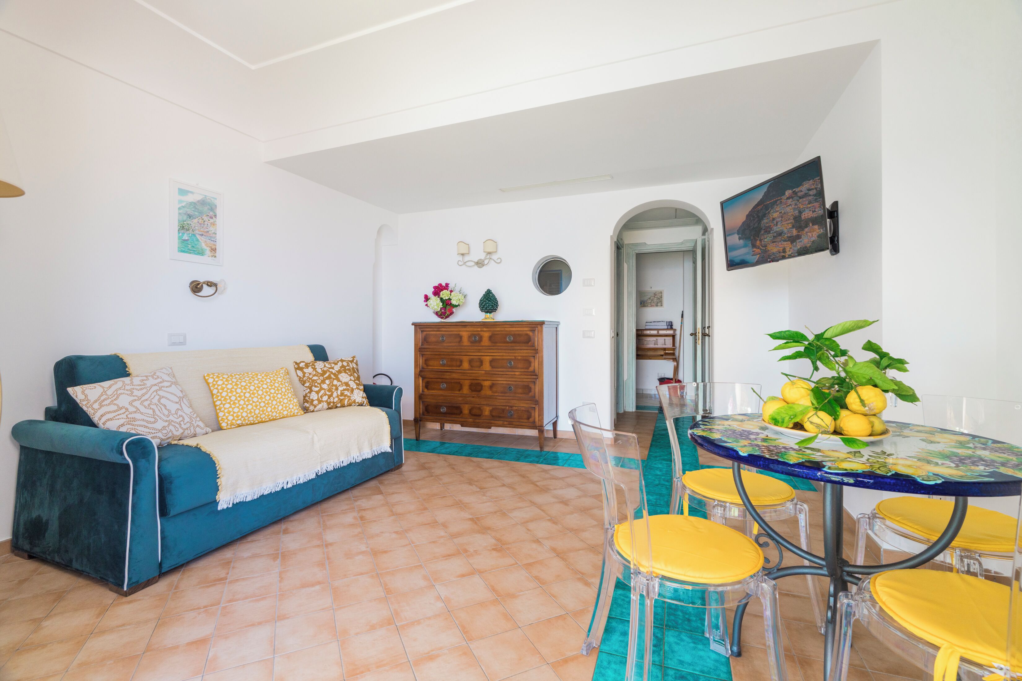 A living room with a blue sofa, a wooden dresser, a glass table with yellow chairs, and a view of a coastal town through an archway.