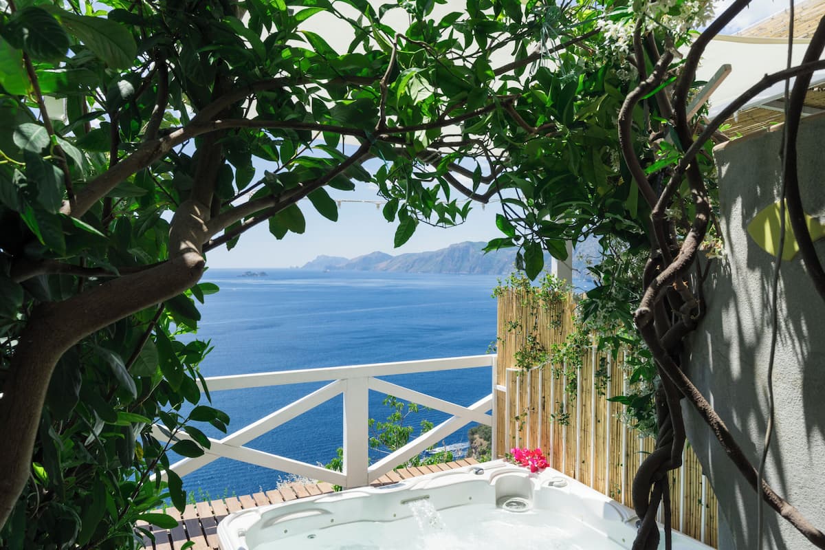 A hot tub with a view of the sea, surrounded by greenery and a wooden fence.