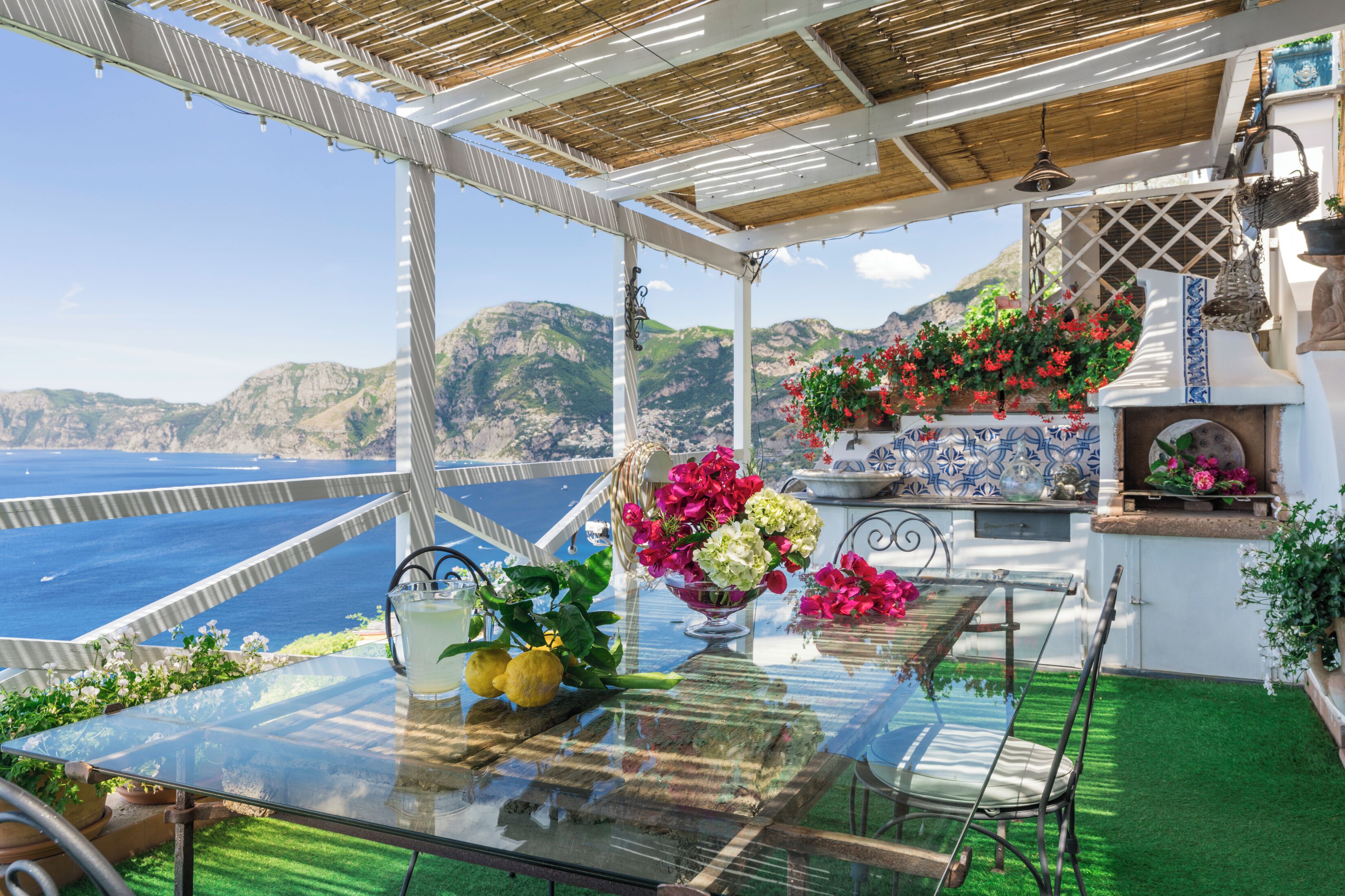 Mesa de vidrio con vista al mar y a las montañas, pared de azulejos blancos y una maceta colgante de flores.
