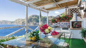 Mesa de vidrio con vista al mar y a las montañas, pared de azulejos blancos y una maceta colgante de flores.