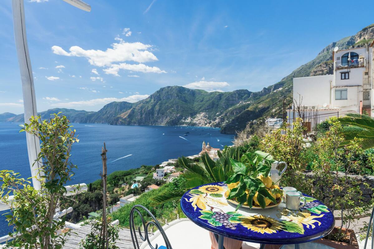 A terrace with a table set for two, overlooking a coastal landscape with mountains and the sea.
