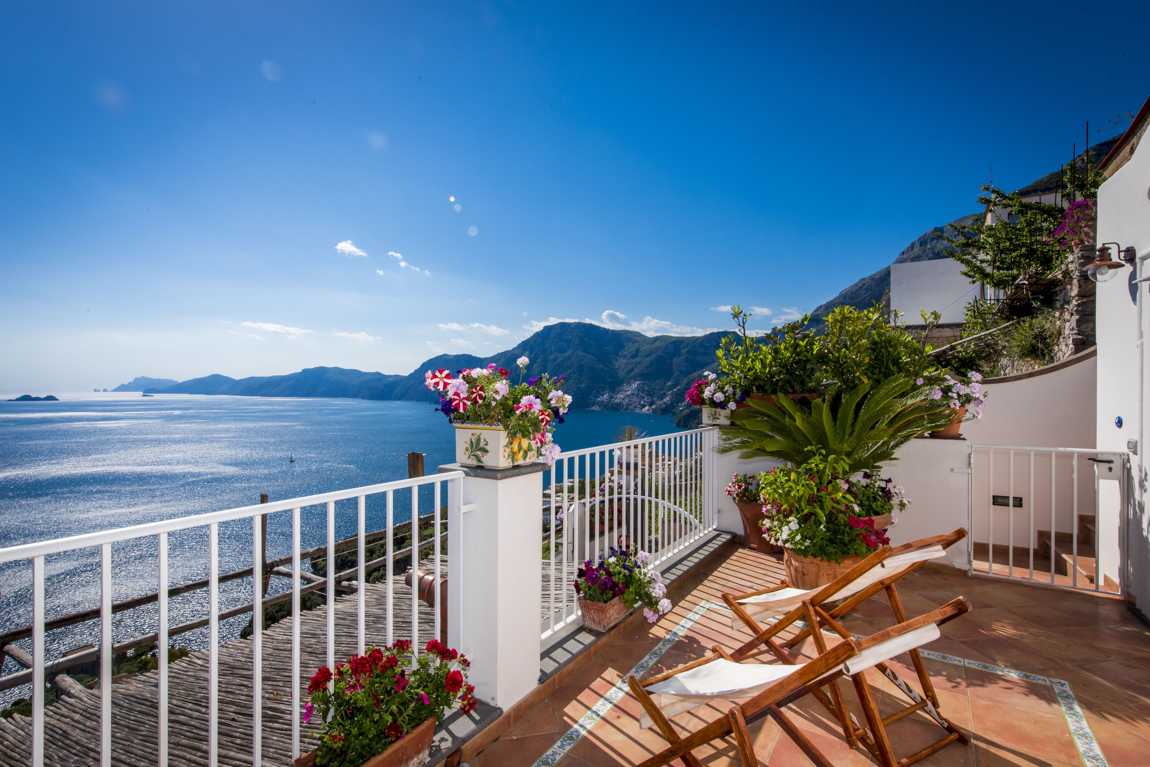 A terrace with a view of the sea, wooden chairs, potted plants, and a white railing.