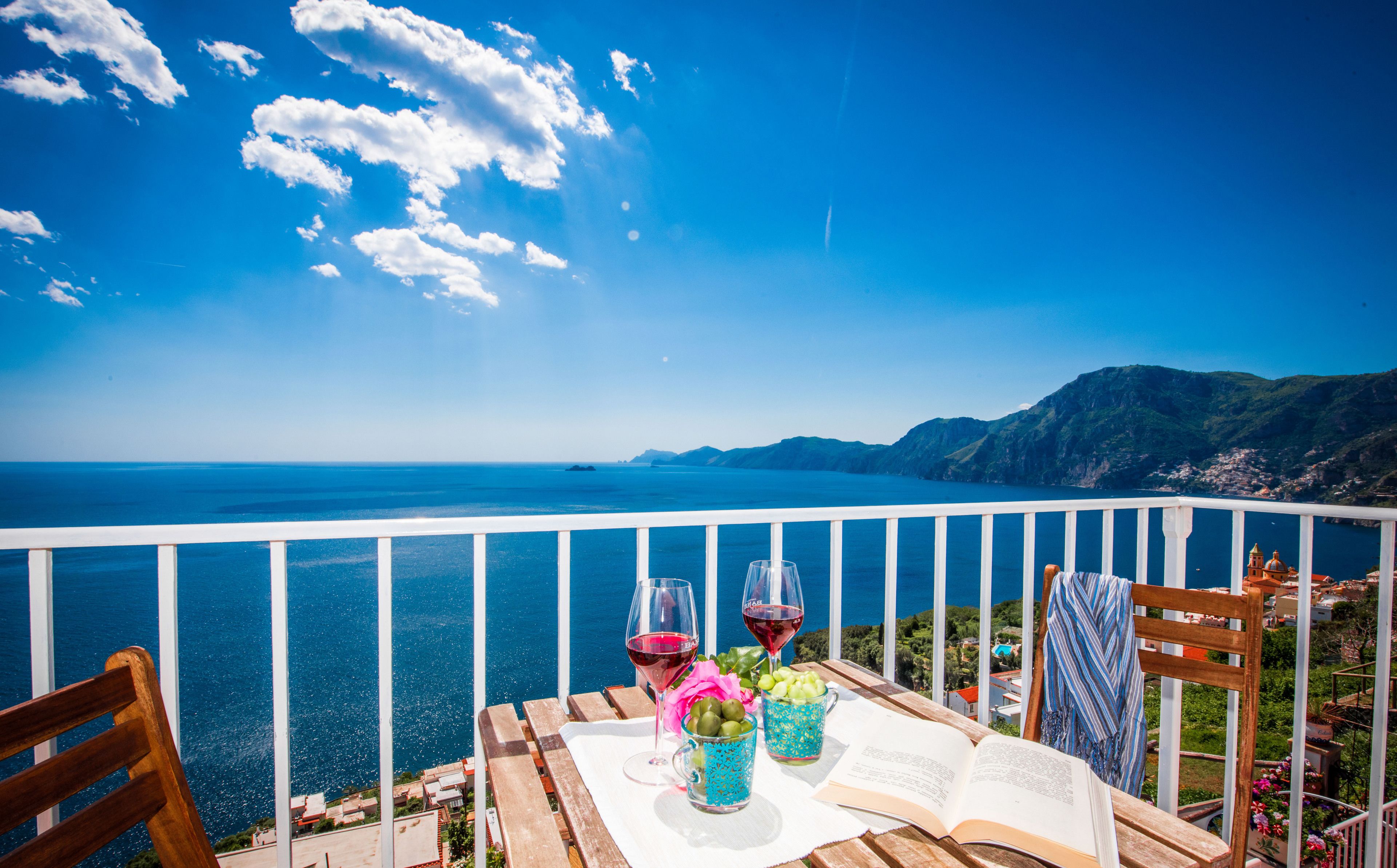 Un balcón con vista al mar, una mesa con un libro, dos copas de vino tinto y una bebida azul y verde.