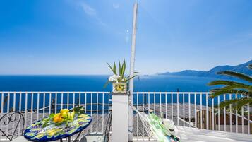 Un balcón con una mesa lista para comer, una silla y una hamaca. Una planta en maceta con flores está sobre un pedestal. Se ve el mar y montañas a lo lejos.
